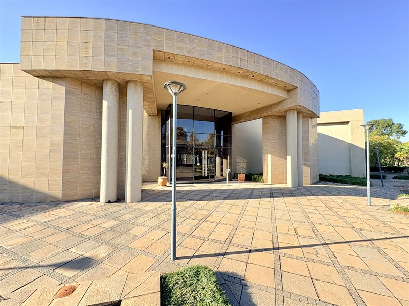 Modern office building entrance featuring a curved stone facade, glass curtain wall, and expansive paved forecourt with integrated lighting.