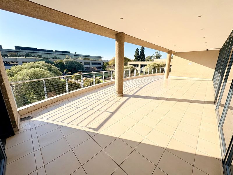 A spacious, covered balcony featuring tiled flooring, recessed lighting, and a metal balustrade, offering an open view of the surrounding office park landscape.