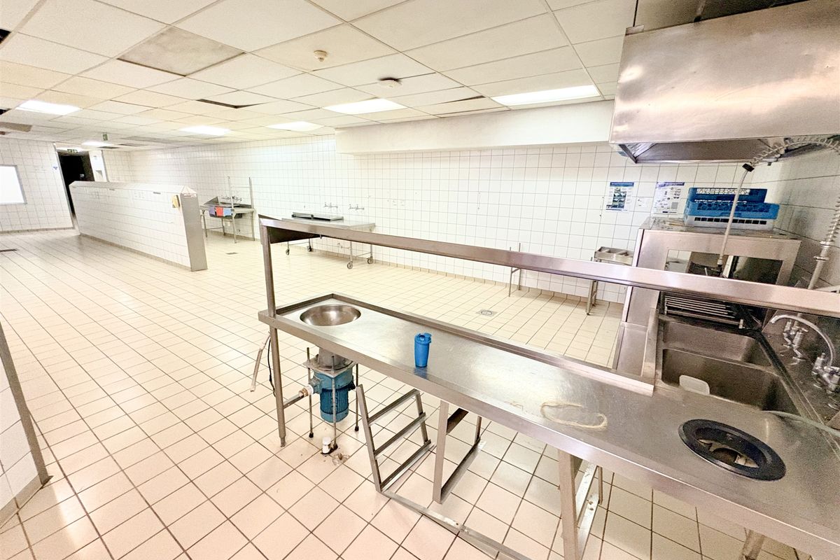 Commercial kitchen space featuring stainless steel prep stations, industrial sinks, extractor hood, tiled surfaces, and suspended ceiling grid.