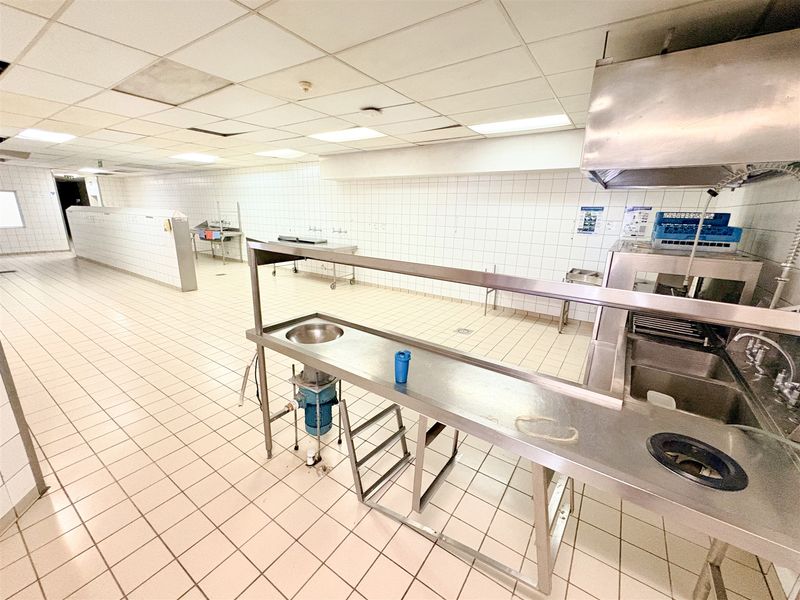 Commercial kitchen space featuring stainless steel prep stations, industrial sinks, extractor hood, tiled surfaces, and suspended ceiling grid.
