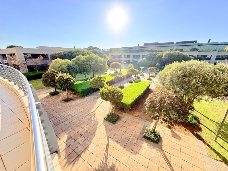 A well-maintained outdoor courtyard and landscaped garden area viewed from an elevated balcony, featuring paved walkways and mature trees.