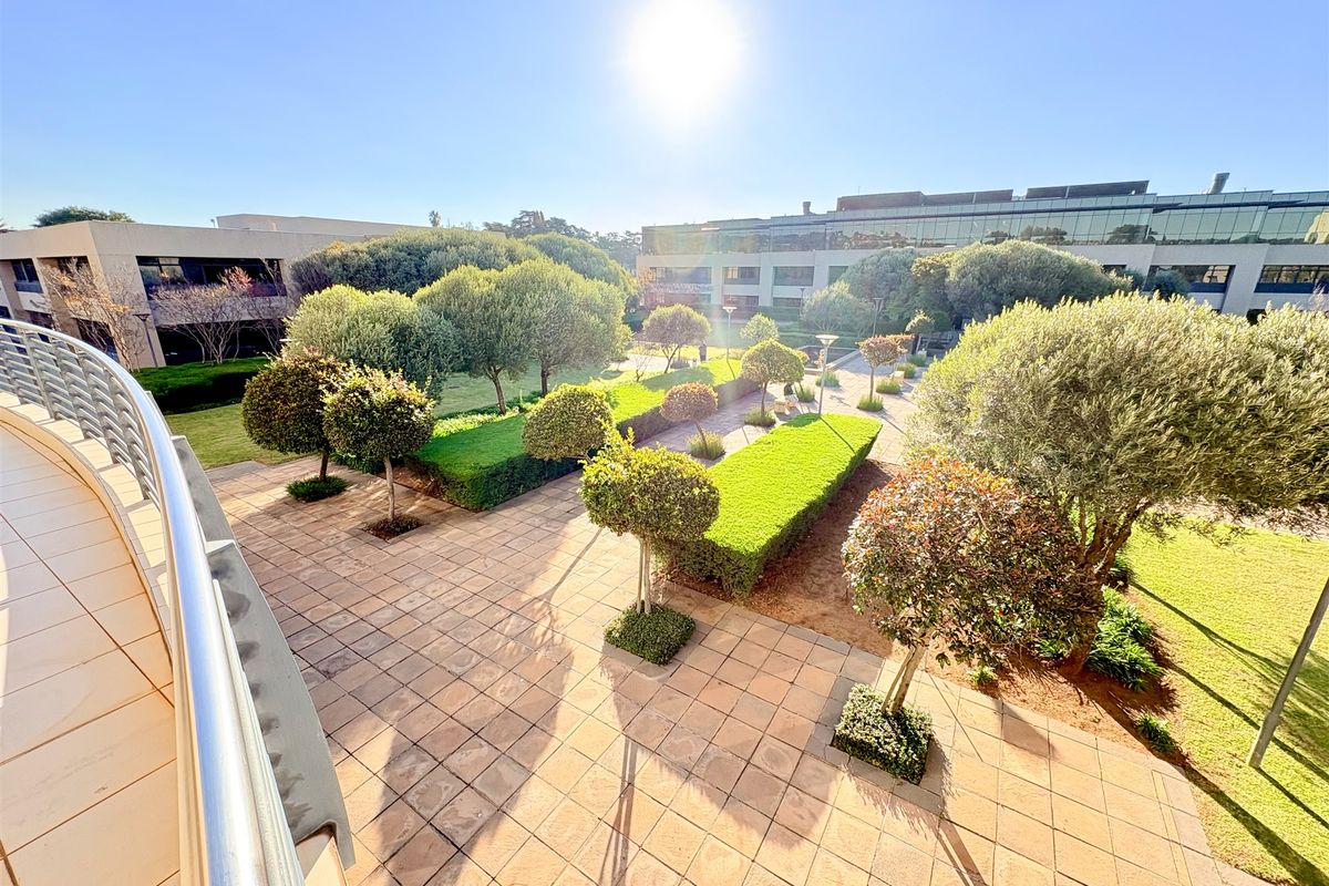 A well-maintained outdoor courtyard and landscaped garden area viewed from an elevated balcony, featuring paved walkways and mature trees.