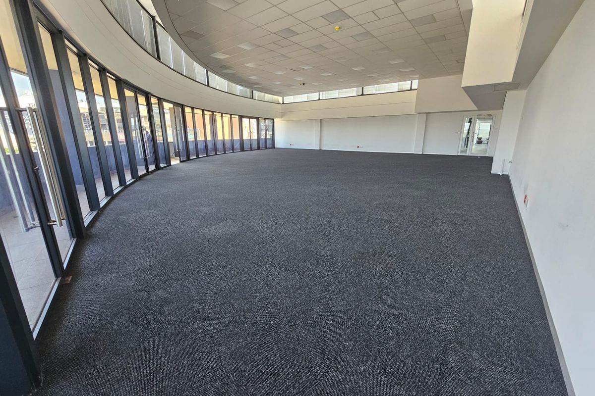 Spacious, open-plan office area featuring curved floor-to-ceiling windows, suspended ceiling tiles, and neutral carpet flooring, offering a bright and professional workspace.