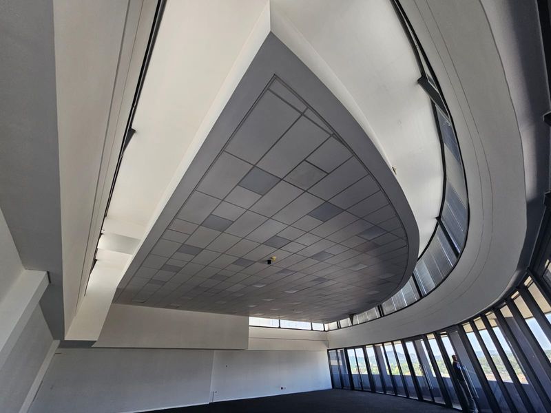 A spacious, modern office area featuring a unique curved suspended ceiling and extensive floor-to-ceiling windows providing ample natural light and panoramic views.
