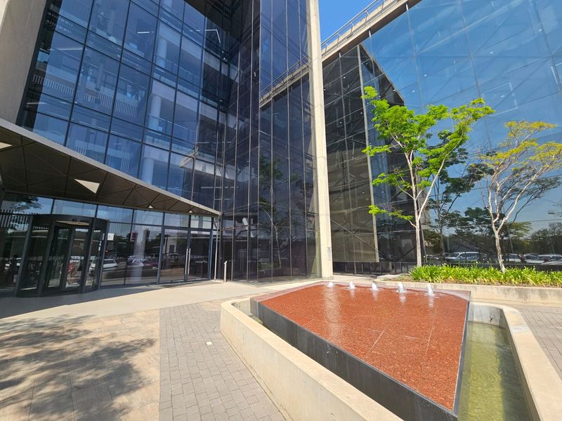 Modern office building entrance featuring a glass facade, a prominent water feature, and a paved pedestrian approach.