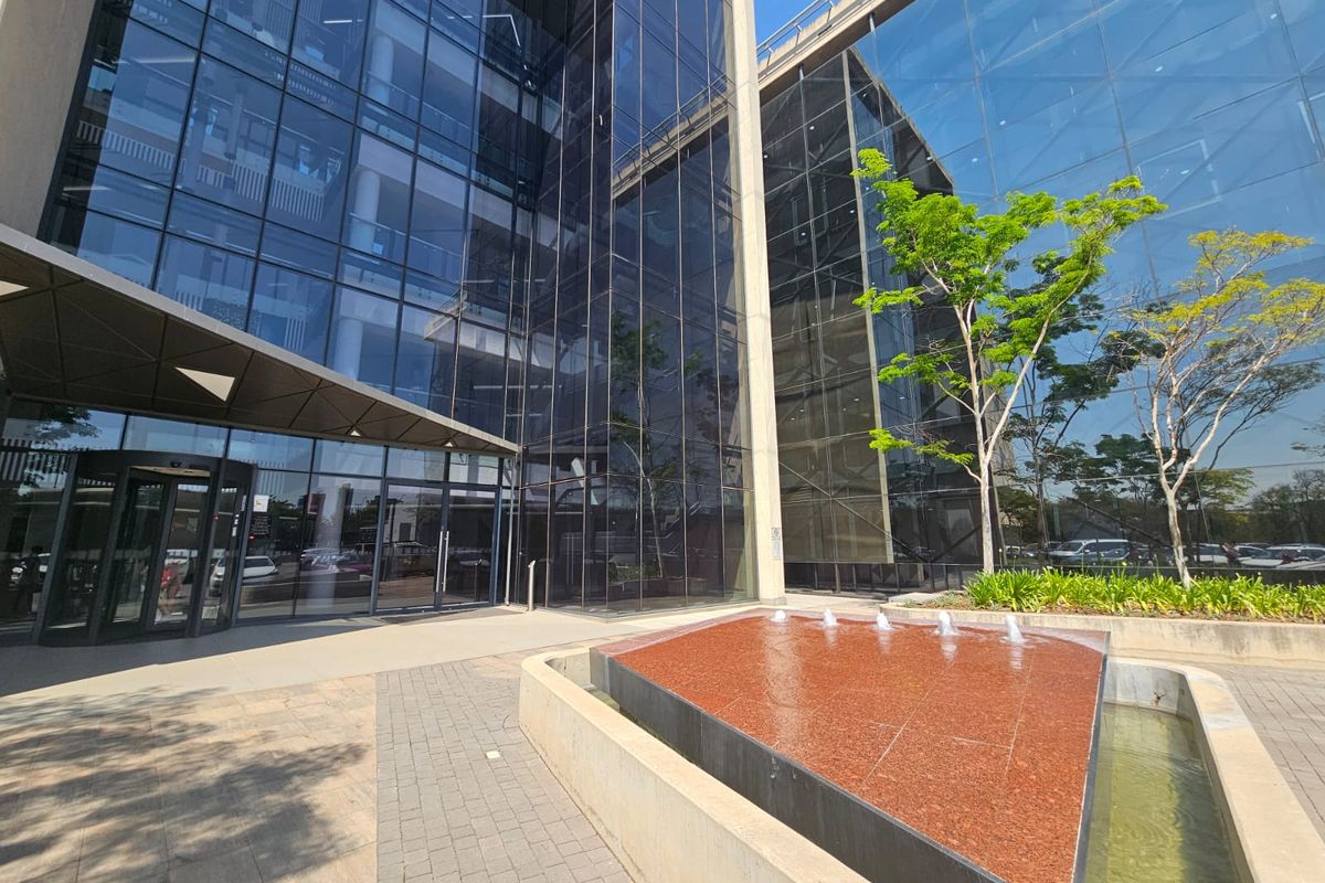 Modern office building entrance featuring a glass facade, a prominent water feature, and a paved pedestrian approach.