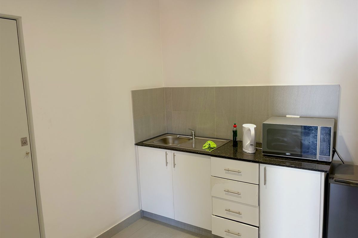 A compact kitchenette featuring white cabinetry, a stainless steel sink, granite-style countertop, and tiled splashback in a clean, functional industrial space.