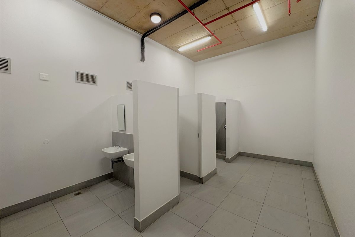 Industrial-style communal bathroom featuring tiled flooring, exposed concrete ceiling with fire sprinkler piping, and multiple partitioned toilet and sink cubicles.