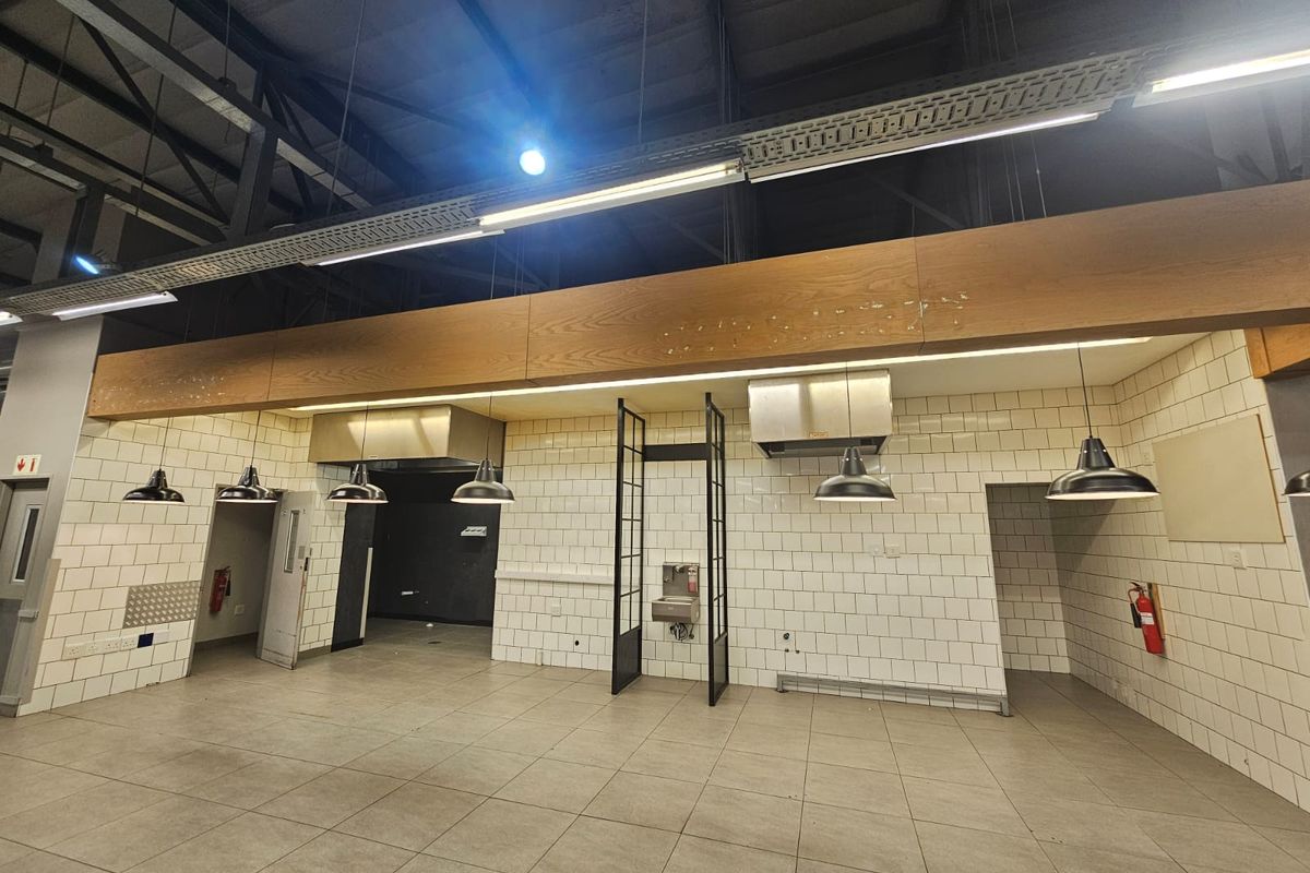 Spacious commercial kitchen area featuring tiled walls, industrial pendant lighting, exposed ceiling structure, and multiple extractor hoods, suitable for a restaurant or food service setup.