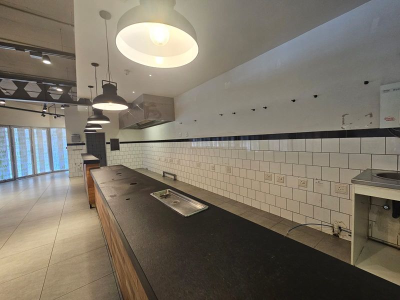 A commercial kitchen space featuring a long built-in counter, subway tile backsplash, extractor fan, and industrial pendant lighting, ready for fit-out.