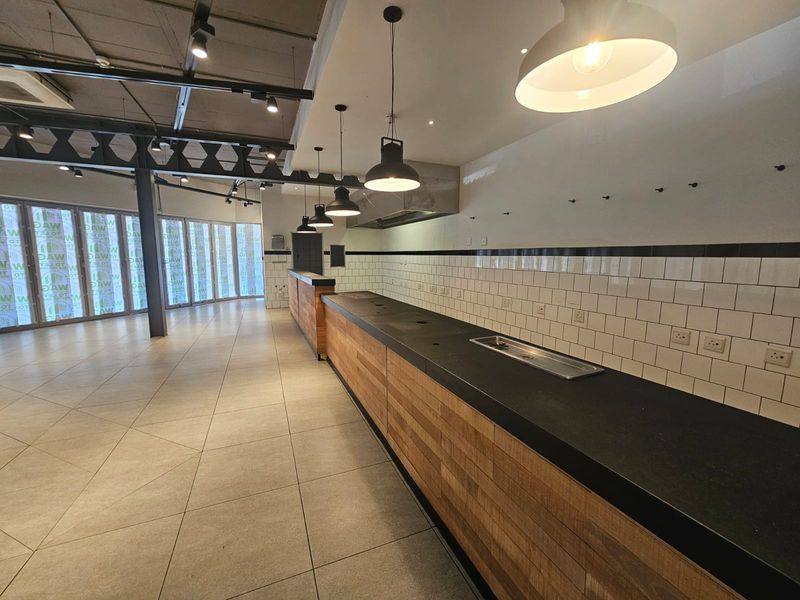 A spacious commercial kitchen area featuring a long service counter, subway tile backsplash, modern pendant lighting, and industrial-style exposed structural elements.