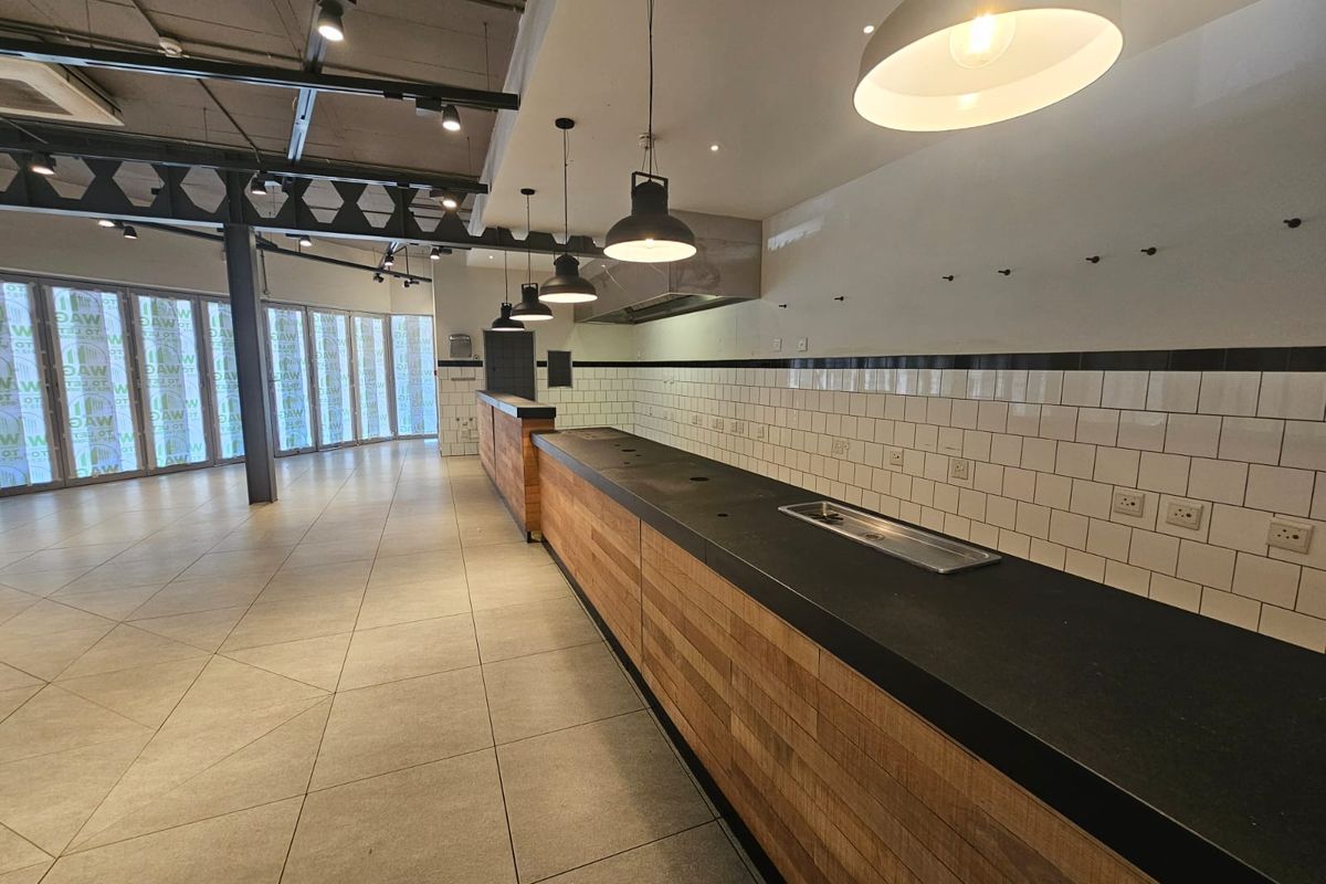 A spacious commercial kitchen area featuring a long service counter, subway tile backsplash, modern pendant lighting, and industrial-style exposed structural elements.