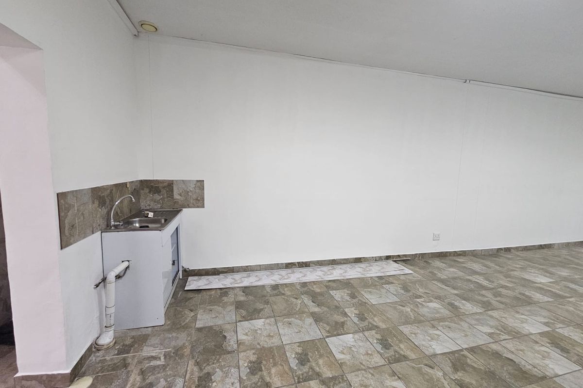 A basic, empty retail kitchenette area featuring tiled flooring, a small stainless steel sink, and exposed plumbing fixtures.