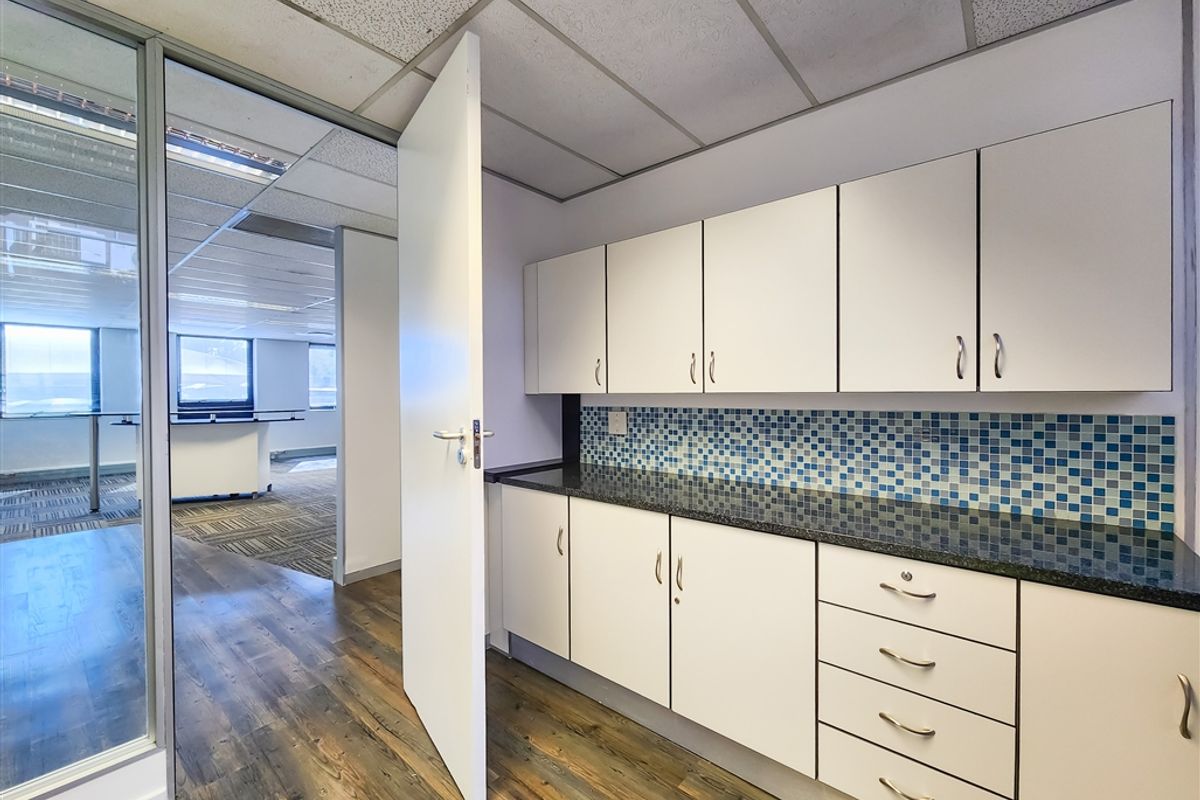 A clean office kitchenette featuring white built-in cabinetry, a dark laminate countertop, and a blue mosaic tile backsplash, adjacent to a glass-partitioned office space.
