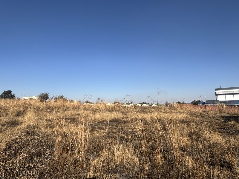 A view of a vacant commercial land plot featuring dry grass and a perimeter brick wall under a clear blue sky.