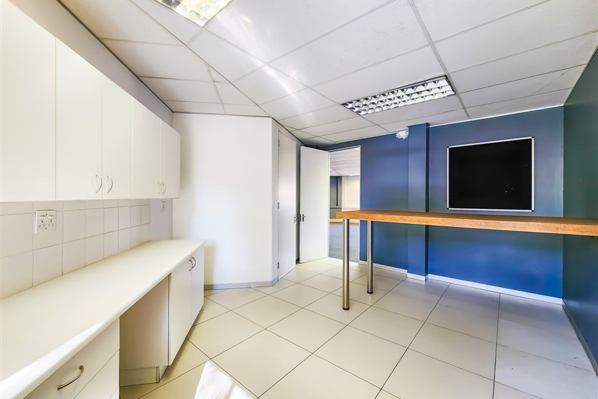 A functional office kitchenette area featuring white cabinetry, tiled flooring, a built-in counter, and a suspended ceiling with integrated lighting.