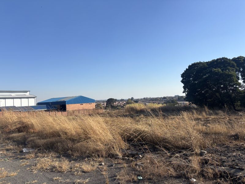 A view of a vacant commercial land plot with dry grass, adjacent to an existing industrial building and perimeter wall.