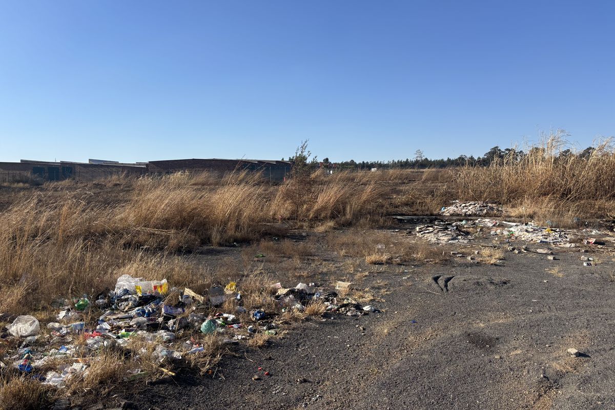 A view of a vacant commercial land plot featuring overgrown dry grass and significant accumulation of litter and debris.