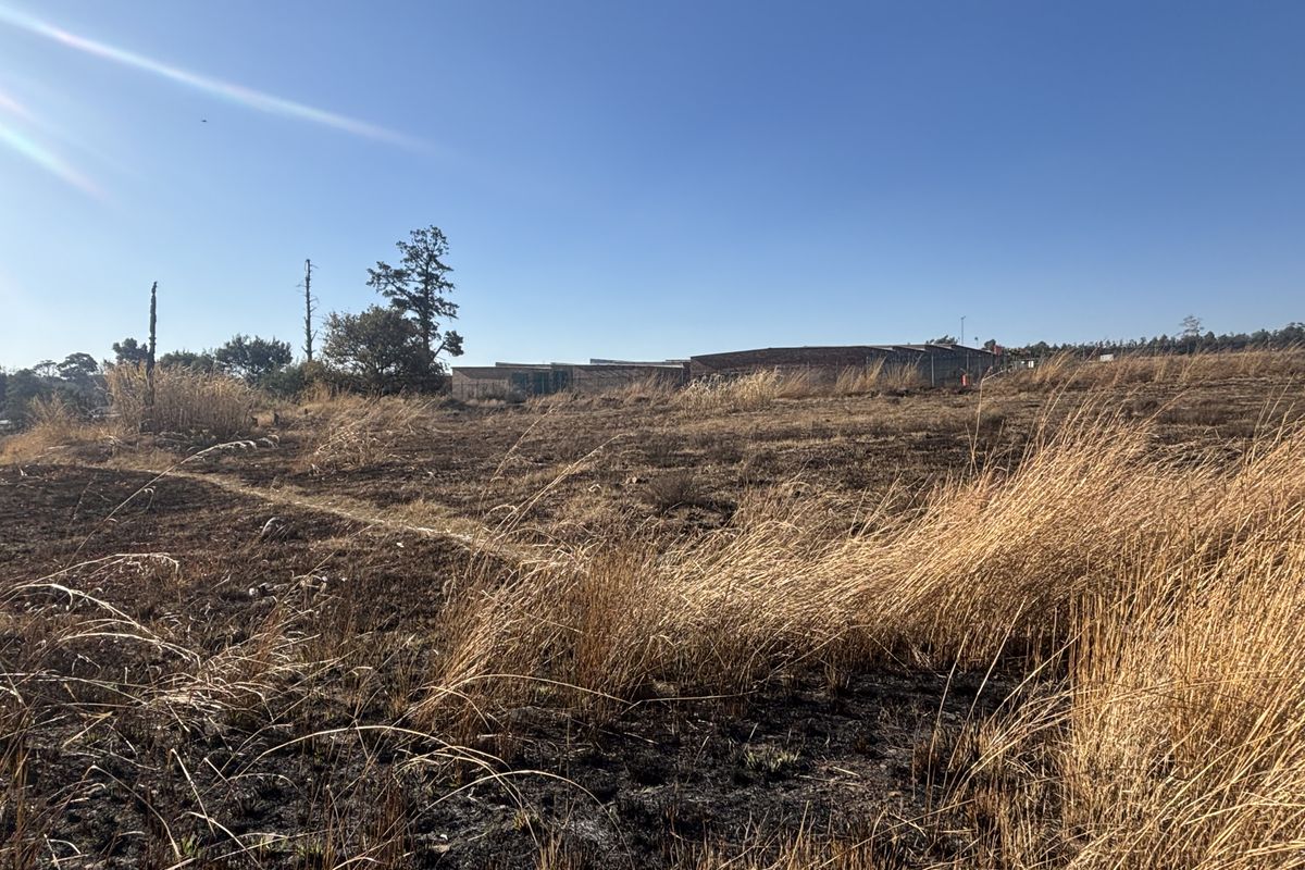 A wide-angle view of a vacant commercial land plot featuring dry grass and open space with distant building structures visible.