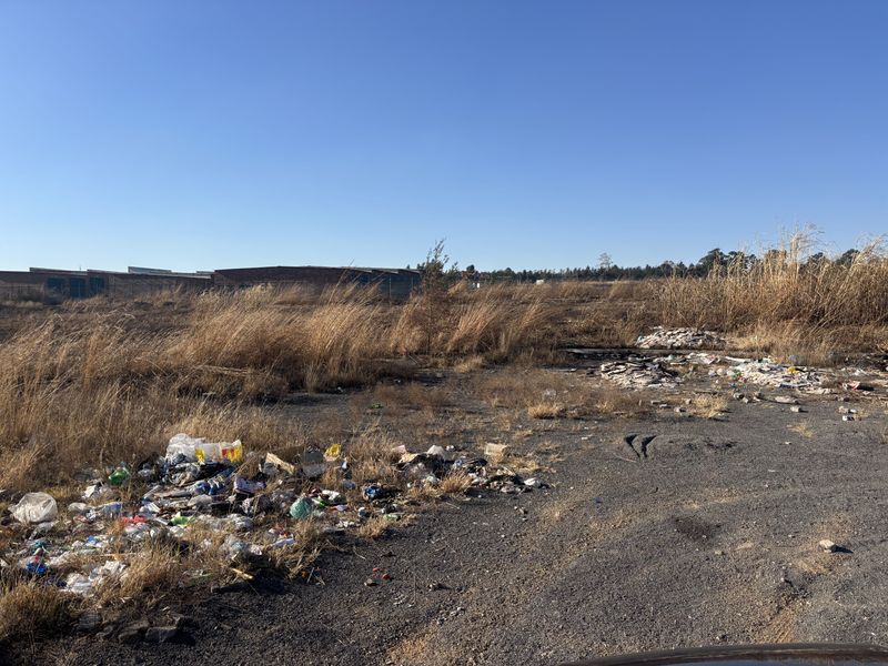 A view of a vacant commercial land plot featuring overgrown dry grass and significant accumulation of litter and debris.