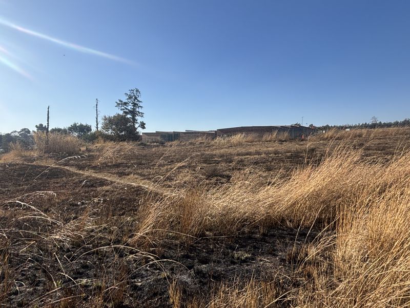 A wide-angle view of a vacant commercial land plot featuring dry grass and open space with distant building structures visible.