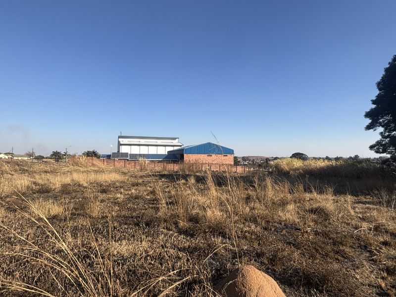 A view of a vacant commercial land plot featuring dry grass, a brick boundary wall, and a large industrial building with a metal roof.