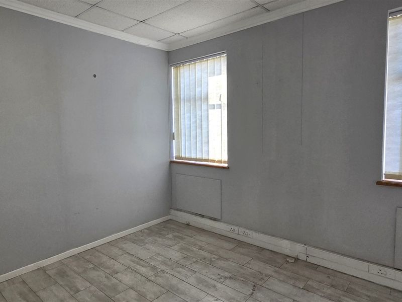 An empty office space featuring wood-look tile flooring, suspended ceiling tiles, and dual windows with vertical blinds.