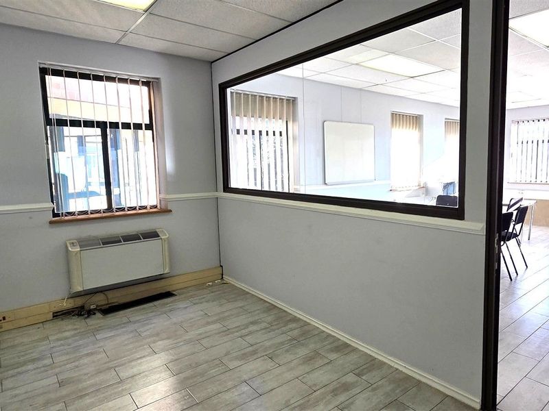 A vacant office space featuring tiled flooring, a glass partition wall, suspended ceiling, and wall-mounted HVAC unit with security bars on the windows.