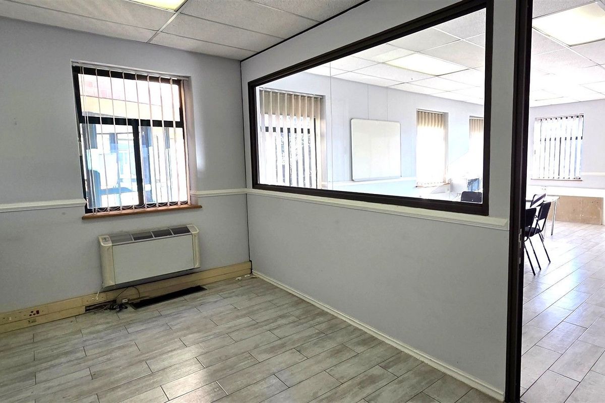 A vacant office space featuring tiled flooring, a glass partition wall, suspended ceiling, and wall-mounted HVAC unit with security bars on the windows.
