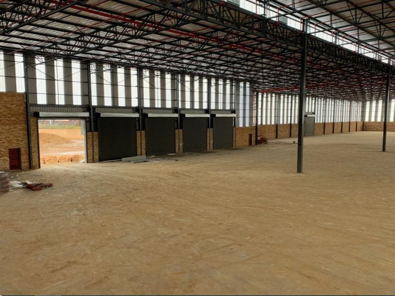 Expansive industrial warehouse space featuring high steel truss ceilings, multiple roller shutter doors, and exposed brick walls, currently in an unfinished shell condition.