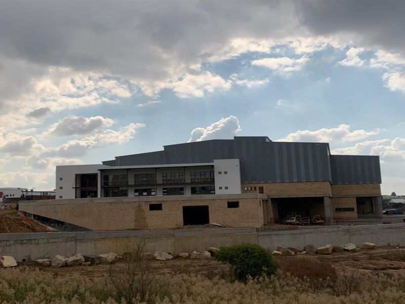 An exterior view of a large industrial building under construction, featuring metal cladding, stone masonry, and a concrete retaining wall.