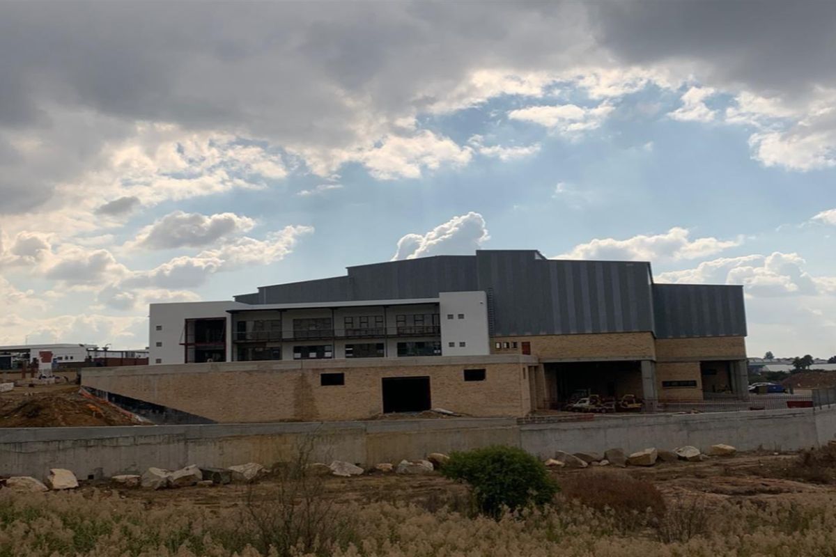 An exterior view of a large industrial building under construction, featuring metal cladding, stone masonry, and a concrete retaining wall.