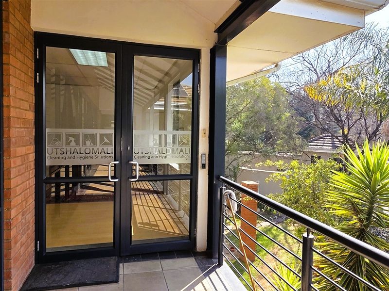 A covered balcony entrance featuring double glass doors, metal railings, and tiled flooring, overlooking a landscaped garden area.
