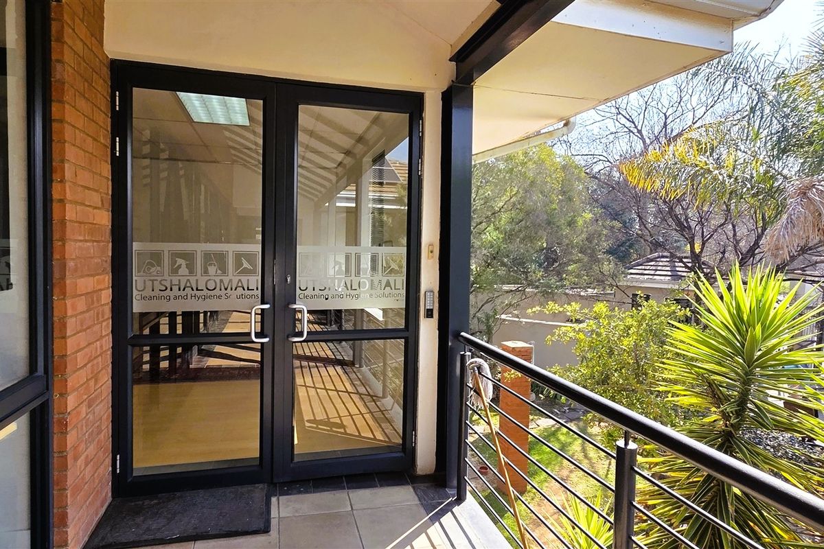 A covered balcony entrance featuring double glass doors, metal railings, and tiled flooring, overlooking a landscaped garden area.