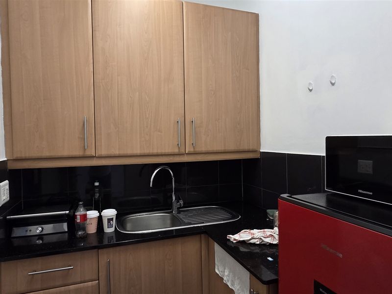 A compact office kitchenette featuring wood-look laminate cabinetry, black granite countertops, a stainless steel sink, and a black tiled backsplash.