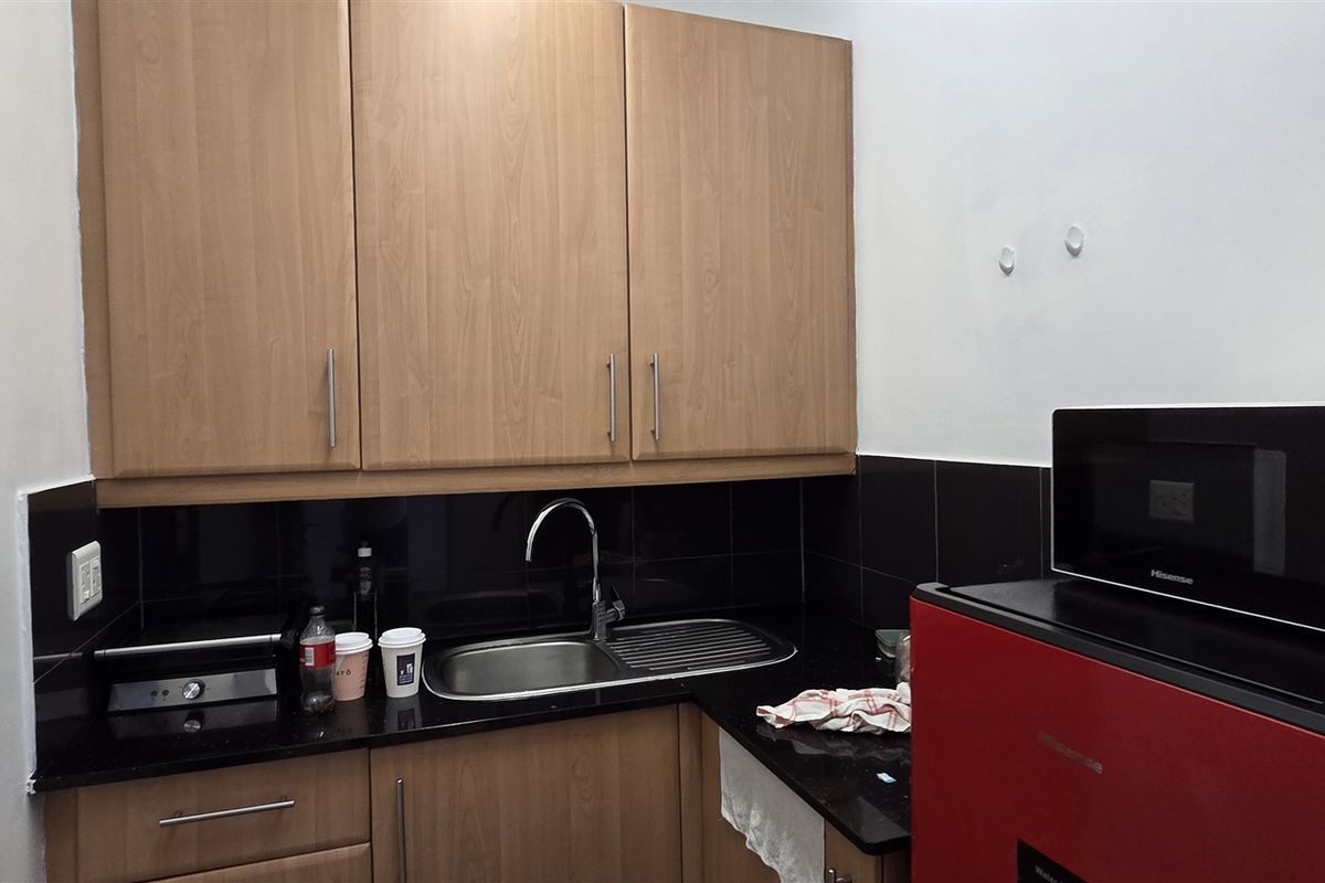 A compact office kitchenette featuring wood-look laminate cabinetry, black granite countertops, a stainless steel sink, and a black tiled backsplash.