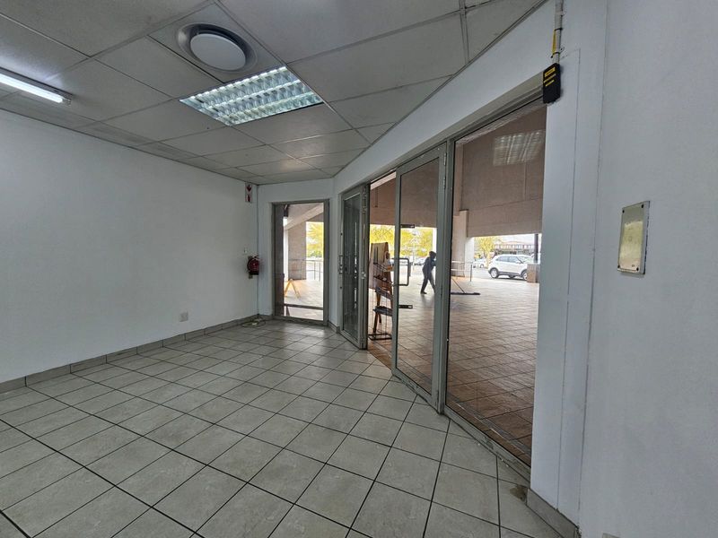 Empty retail entrance area featuring tiled flooring, suspended ceiling with recessed lighting, and large aluminium-framed glass doors providing street access.