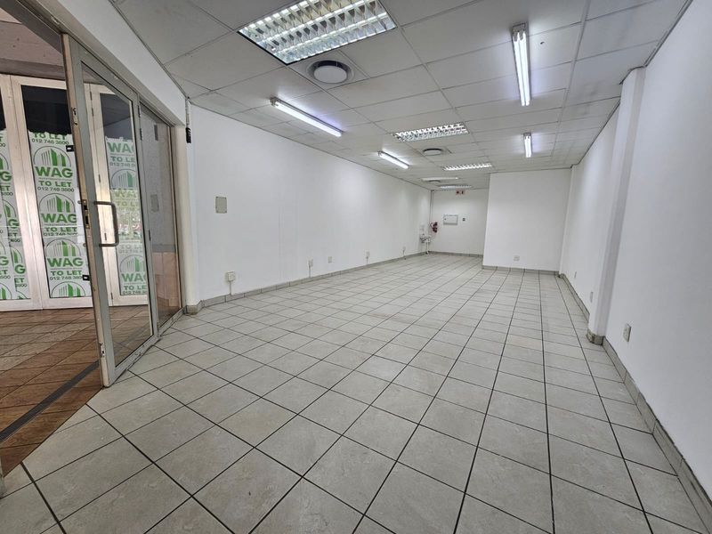 An empty open-plan retail space featuring ceramic tiled flooring, a suspended ceiling with fluorescent lighting, and a glass entrance door.