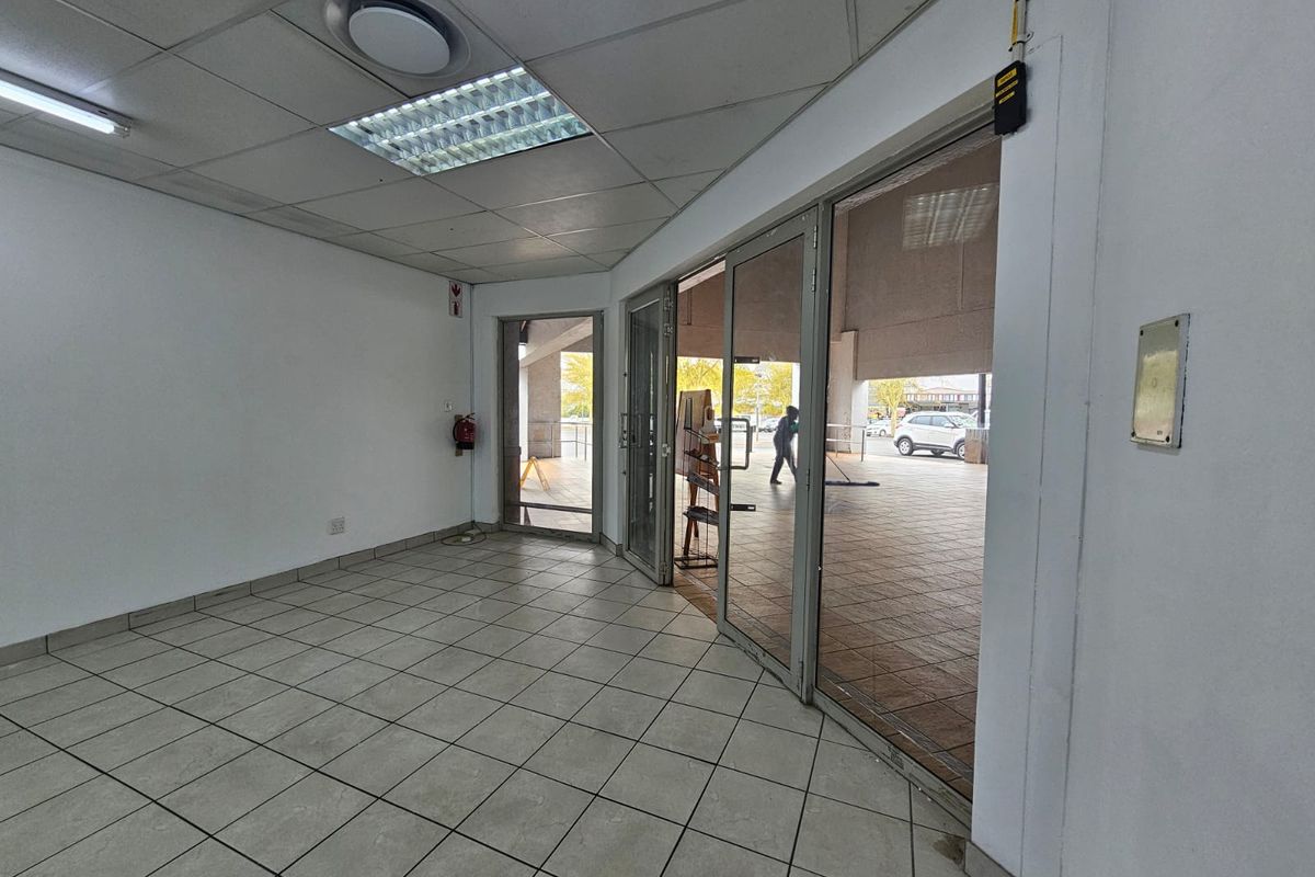 Empty retail entrance area featuring tiled flooring, suspended ceiling with recessed lighting, and large aluminium-framed glass doors providing street access.