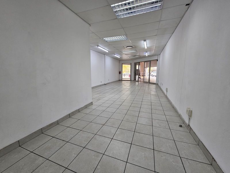 An empty, open-plan retail space featuring ceramic tiled flooring, a suspended ceiling with integrated lighting, and a glass storefront entrance.