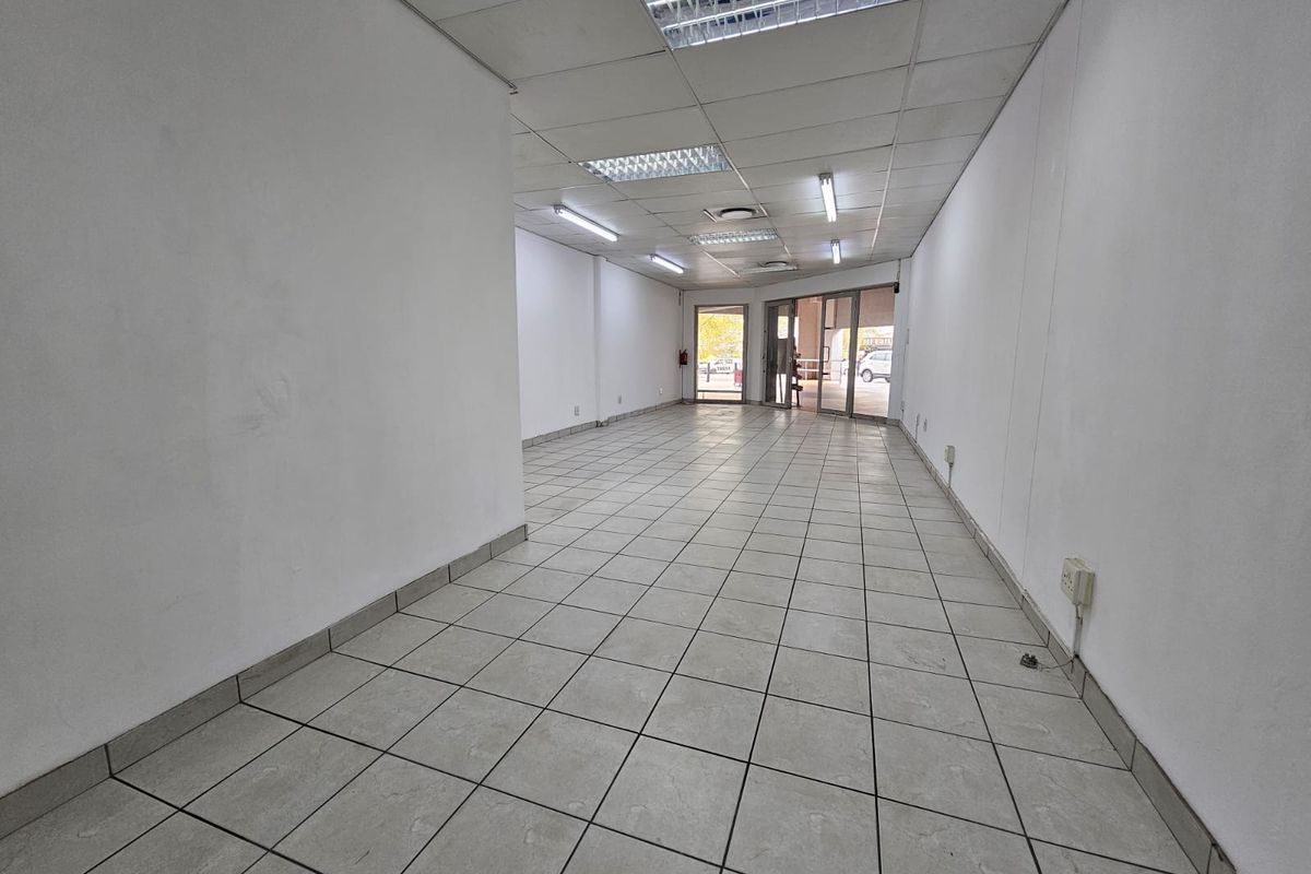 An empty, open-plan retail space featuring ceramic tiled flooring, a suspended ceiling with integrated lighting, and a glass storefront entrance.