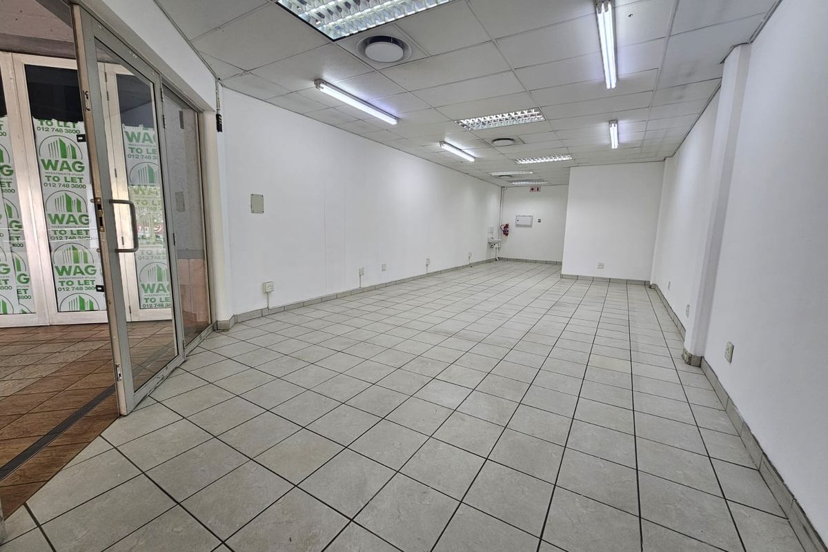An empty open-plan retail space featuring ceramic tiled flooring, a suspended ceiling with fluorescent lighting, and a glass entrance door.