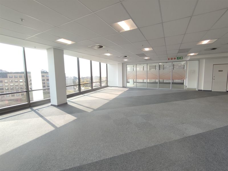 A spacious, vacant open-plan office area featuring large floor-to-ceiling windows, suspended ceilings with recessed lighting, and modern glass partitioning.