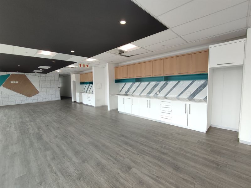 Modern office kitchenette featuring clean white cabinetry, wood-tone upper cupboards, stylish tiled backsplash, and durable laminate flooring in an open-plan workspace.