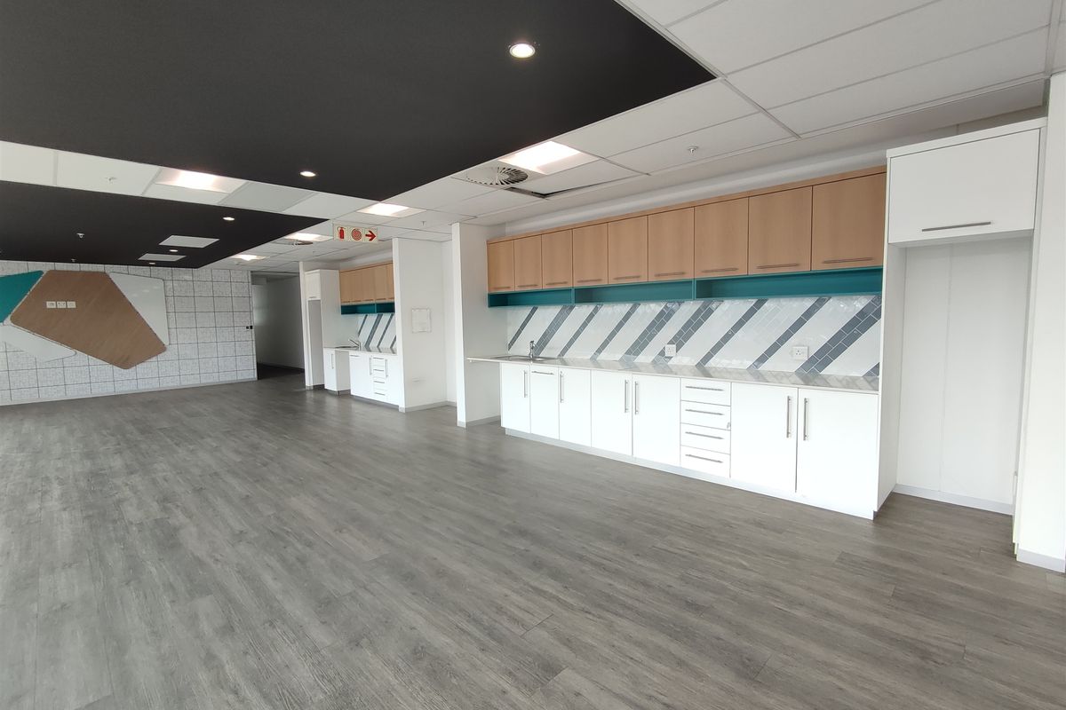 Modern office kitchenette featuring clean white cabinetry, wood-tone upper cupboards, stylish tiled backsplash, and durable laminate flooring in an open-plan workspace.