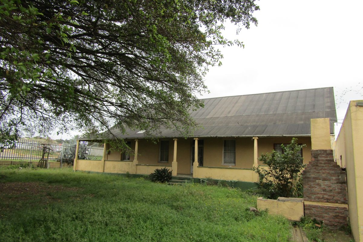 Rear view of a commercial property featuring a covered veranda, corrugated metal roof, and an external brick chimney, set within an overgrown garden area.