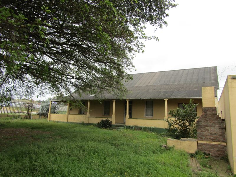 Rear view of a commercial property featuring a covered veranda, corrugated metal roof, and an external brick chimney, set within an overgrown garden area.
