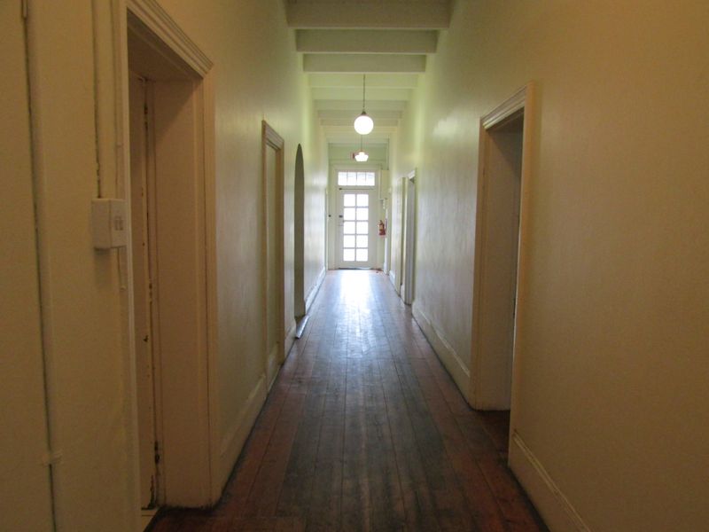 A long, vacant hallway featuring exposed ceiling beams, original hardwood flooring, and a glass-paneled door providing natural light at the end of the corridor.