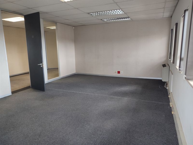 An empty, open-plan office space featuring grey carpeting, suspended ceilings with recessed lighting, and a wall-mounted air conditioning unit.