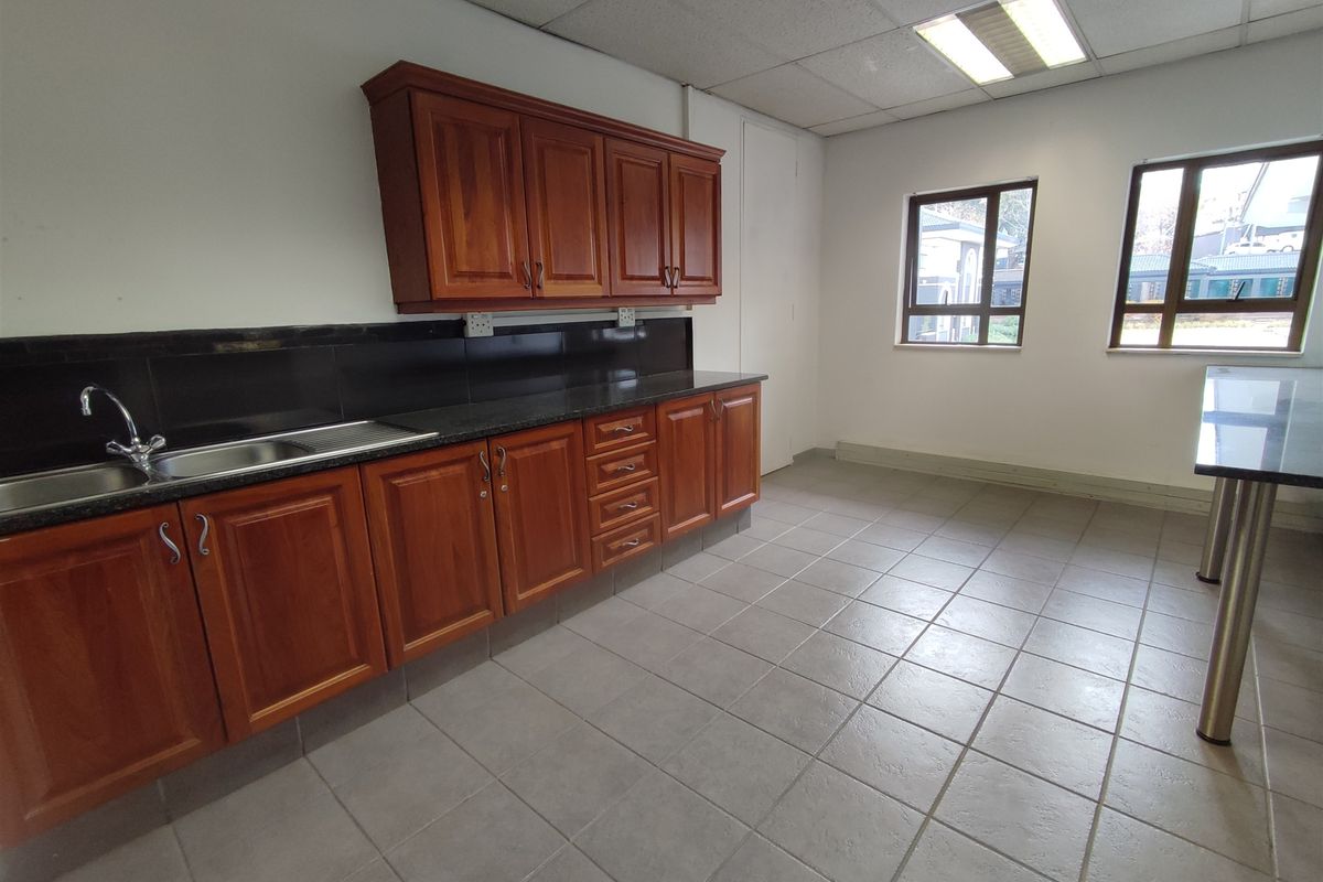 A functional office kitchen area featuring wooden cabinetry, granite countertops, a stainless steel sink, and tiled flooring.